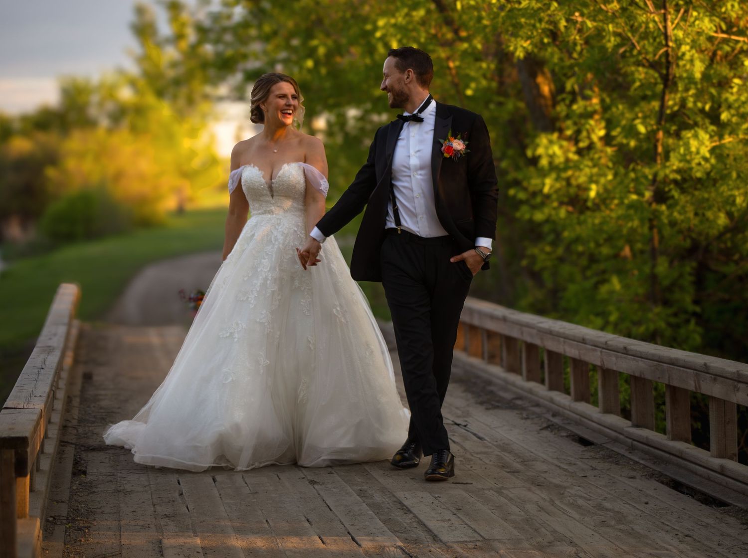 A couple in wedding attire walking together on a wooden bridge path during golden hour.