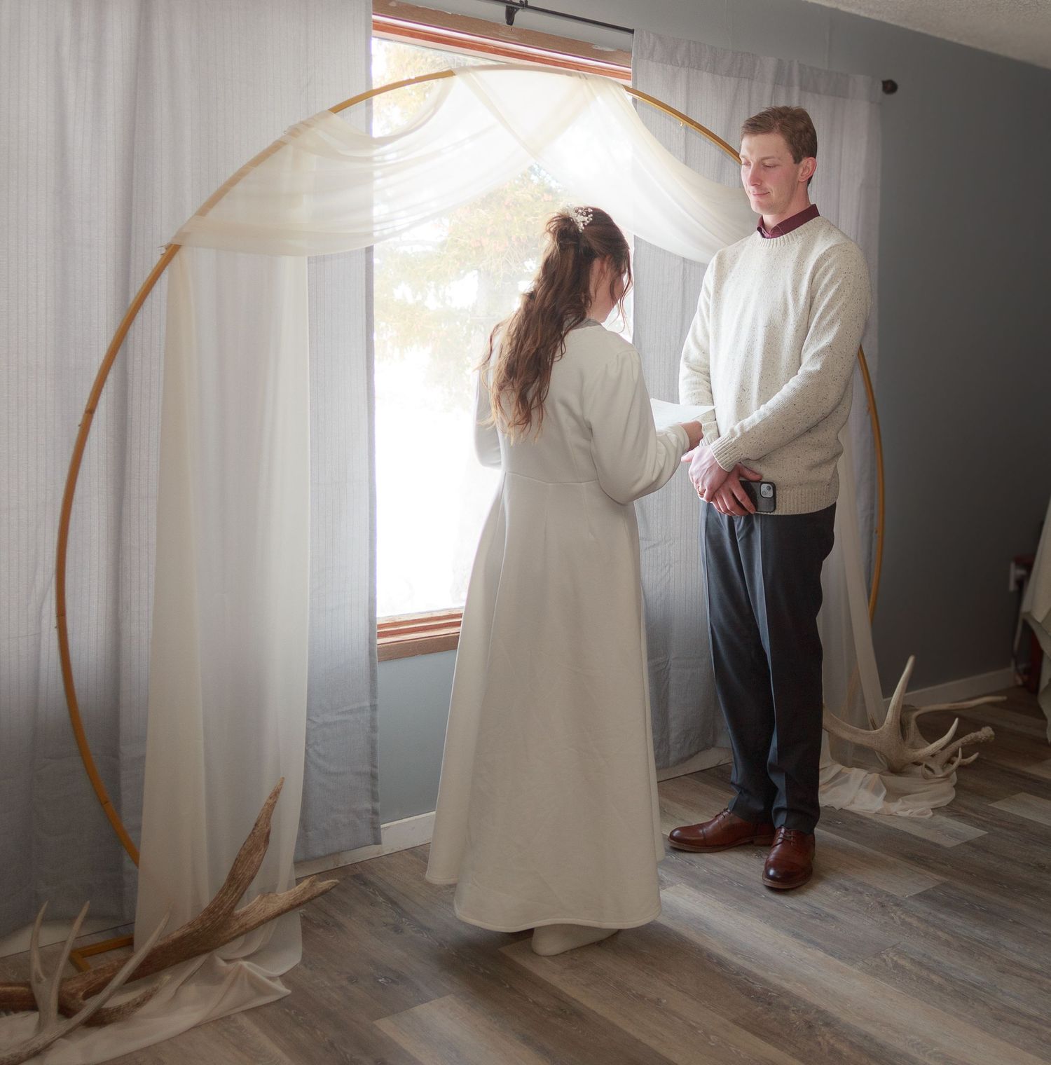 A couple stands together in a minimalist room with white walls and natural lighting.