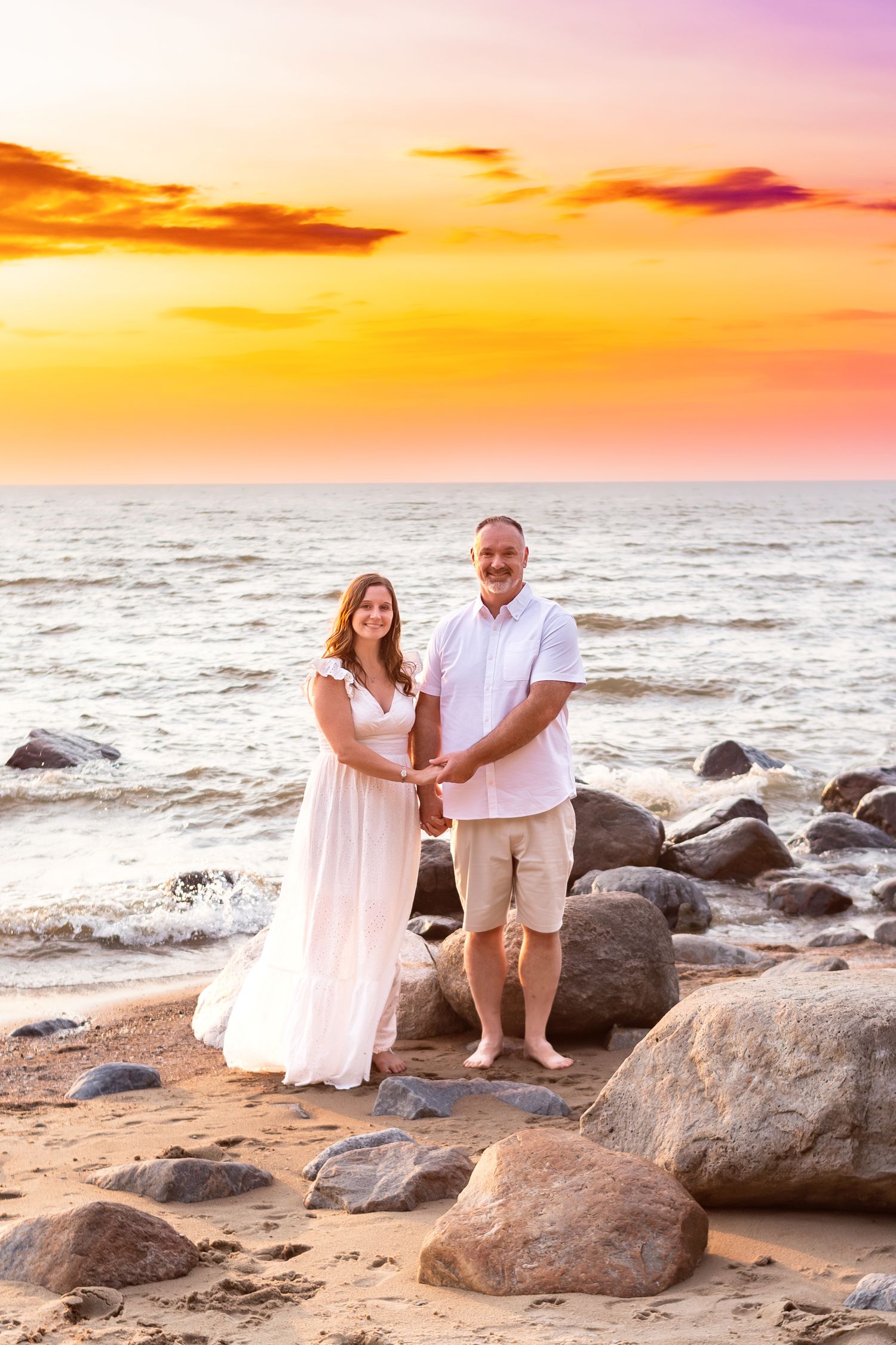 Couple in white attire posing on rocky beach during vibrant sunset.