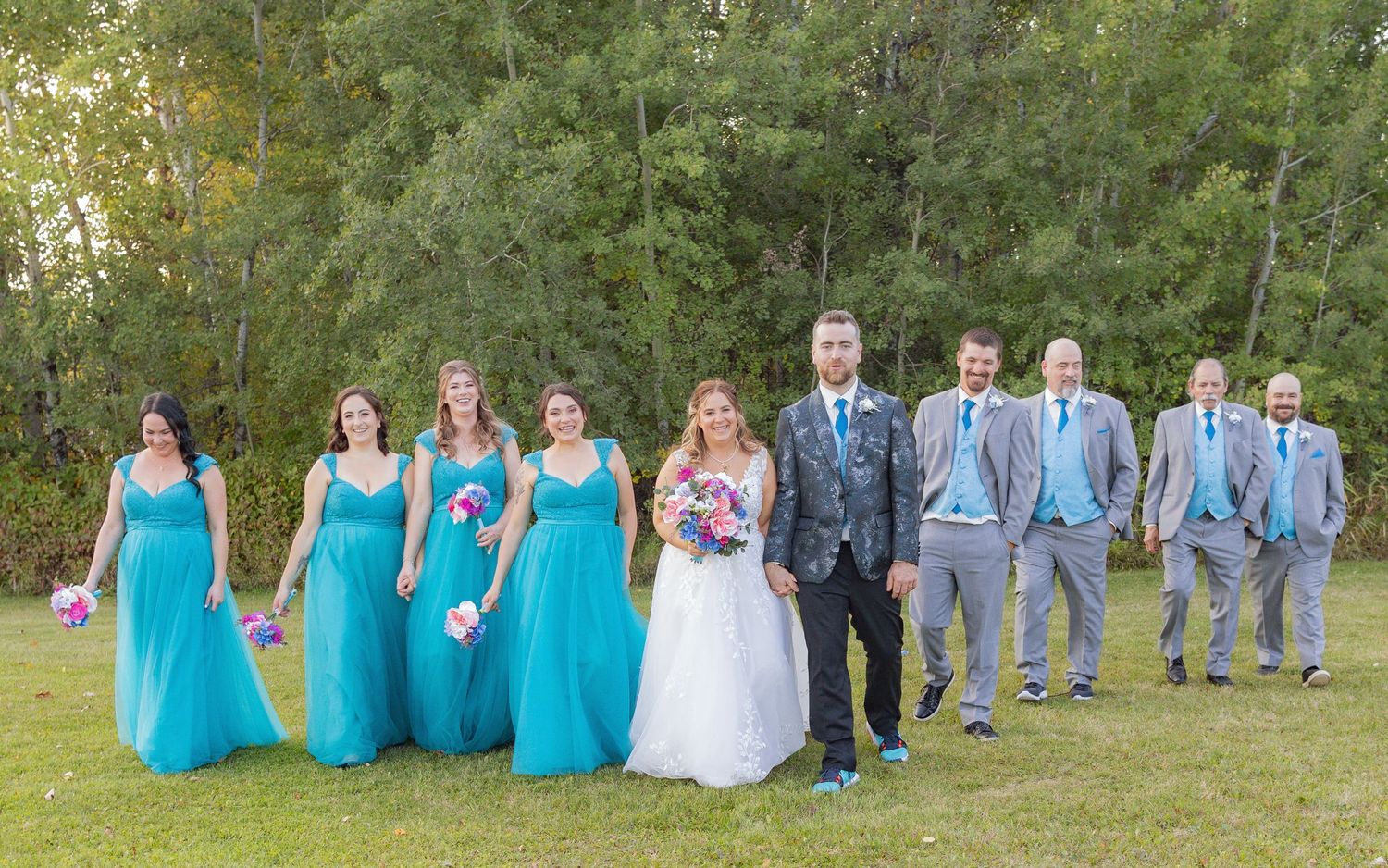Wedding party members lined up on grass wearing coordinated turquoise and gray outfits.