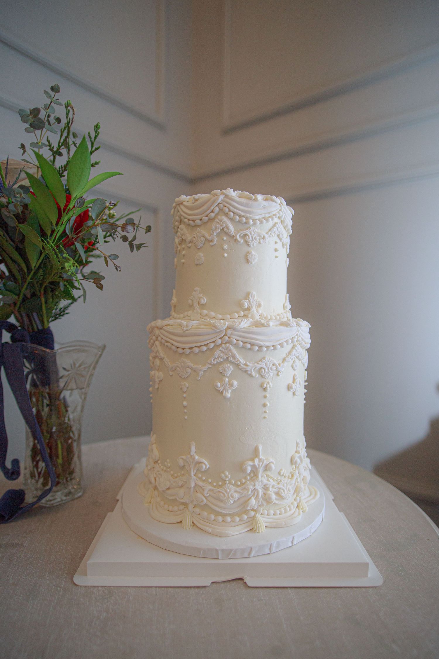 Classic white wedding cake with delicate lace piping and pearl details on a white tablecloth.