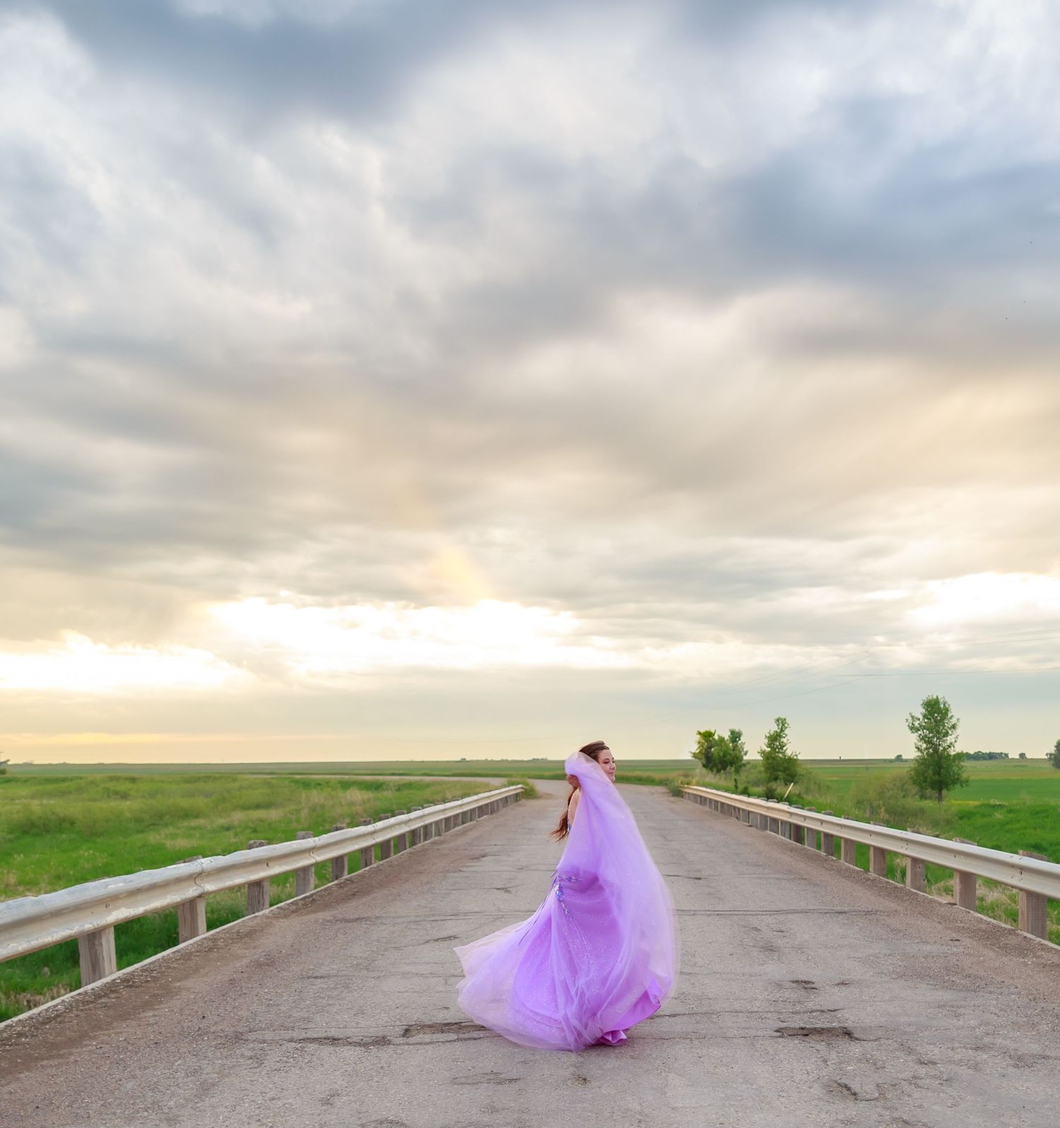 Purple gown billows in the wind on an empty rural road at sunset.