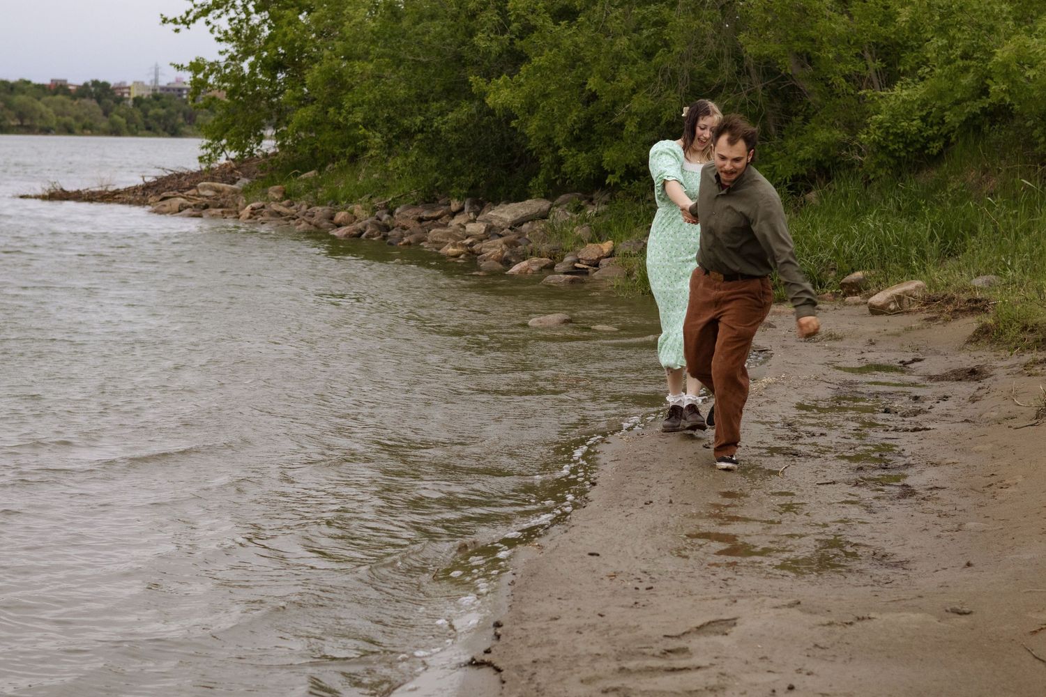 A couple walks together along a rocky riverbank surrounded by nature.