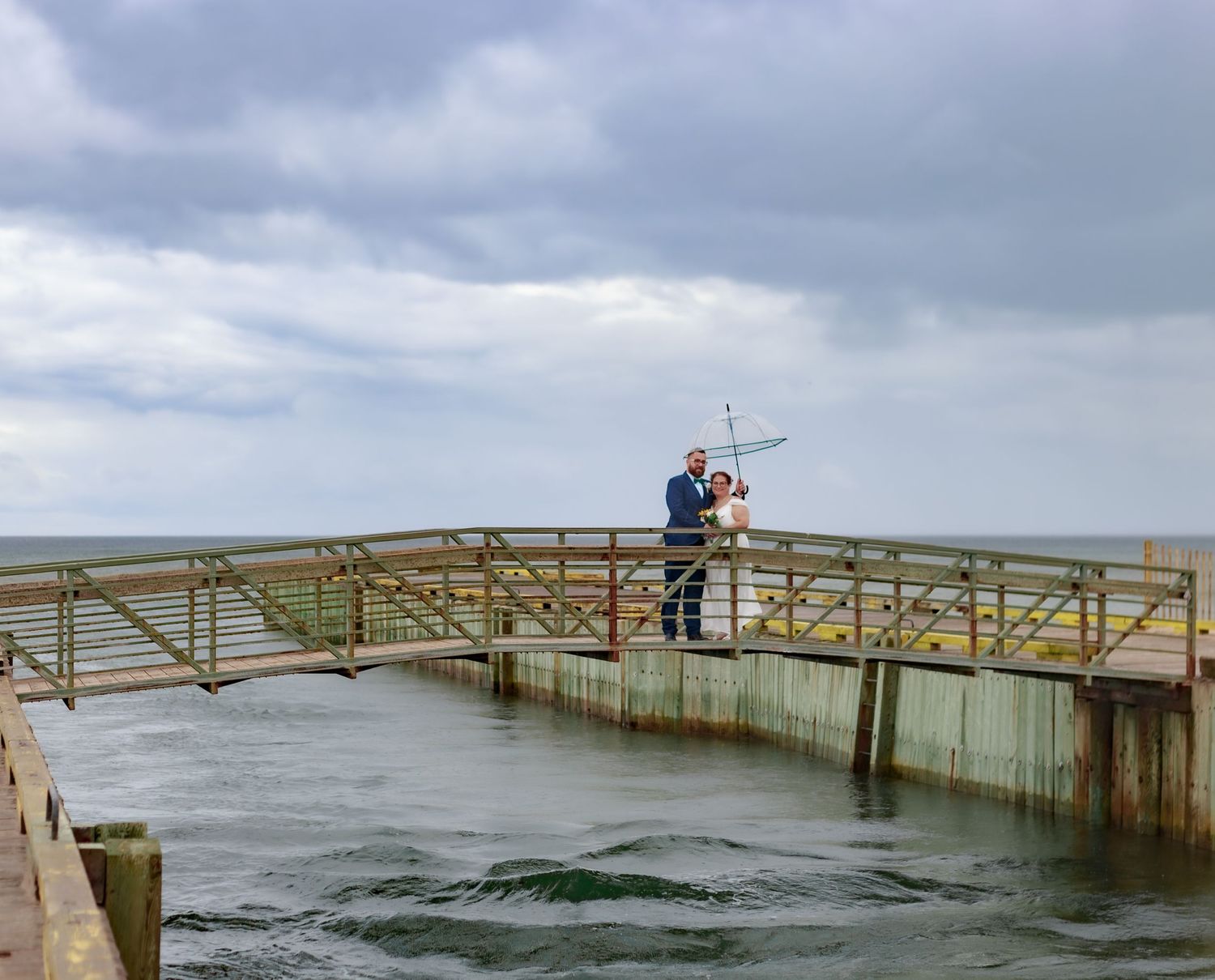 Wedding couple standing on a wooden pier overlooking stormy ocean waters.