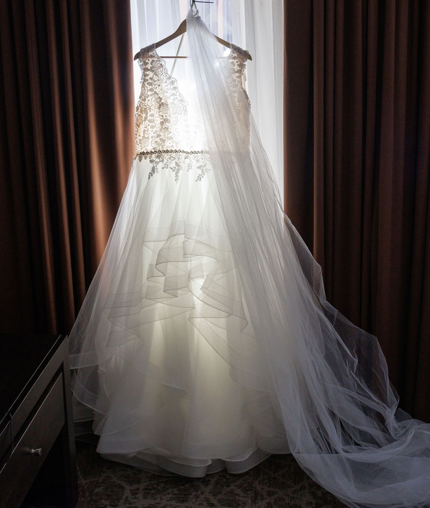 A white wedding dress hanging in silhouette against window light with flowing tulle fabric.