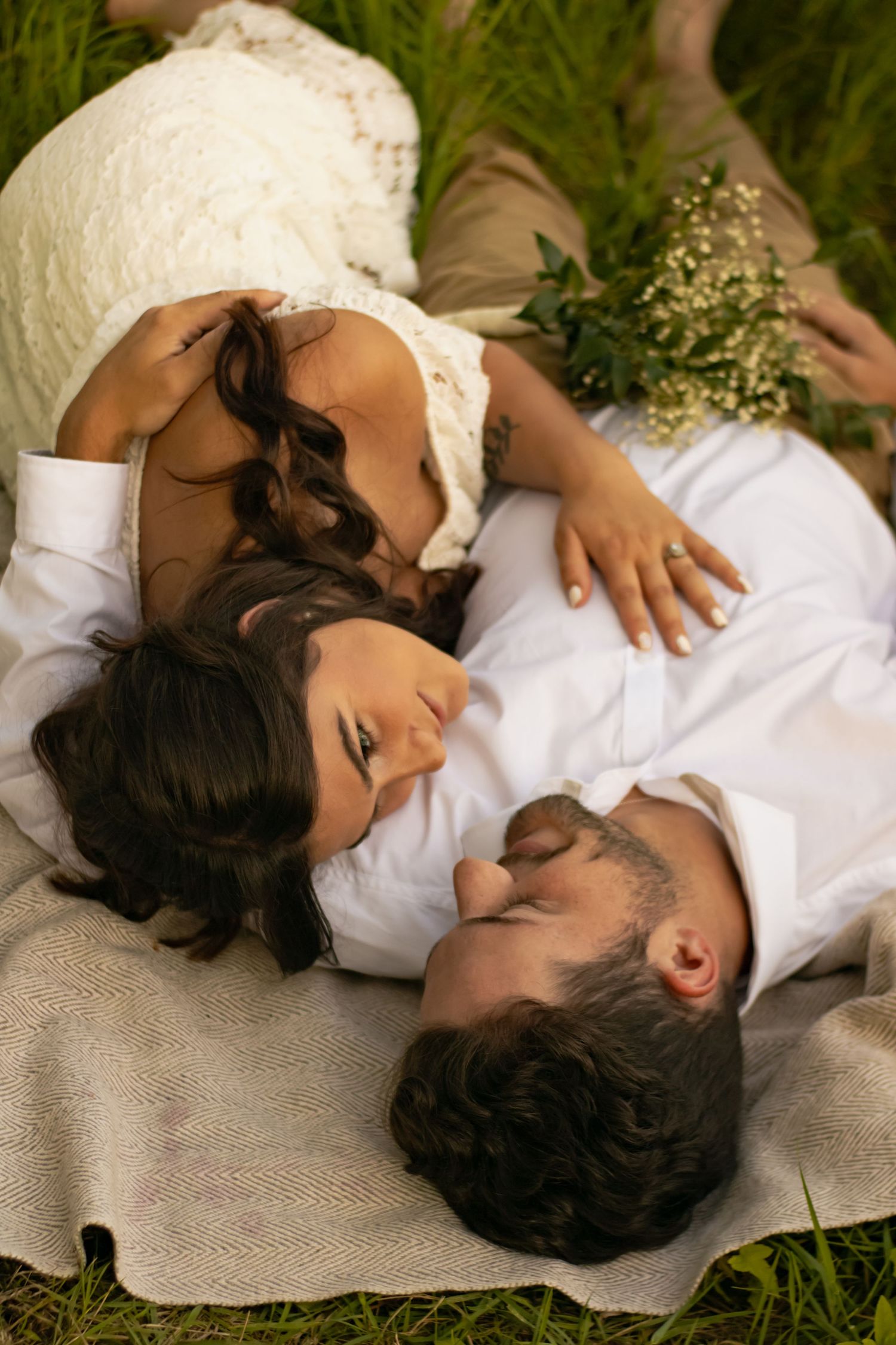 A couple in white attire lying on a blanket in the grass during a romantic photoshoot.