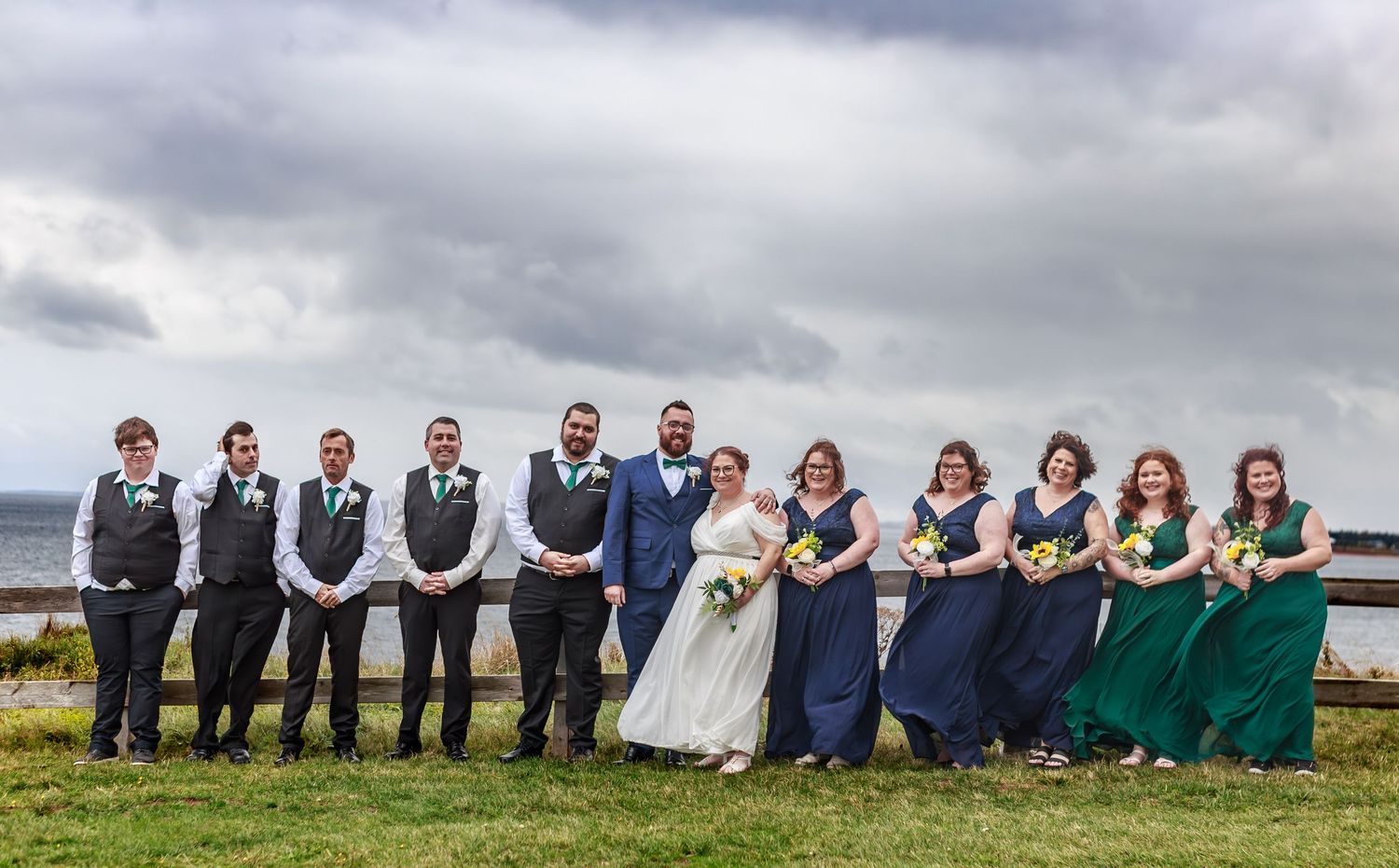 Wedding party in navy and green attire posing along a coastal fence.
