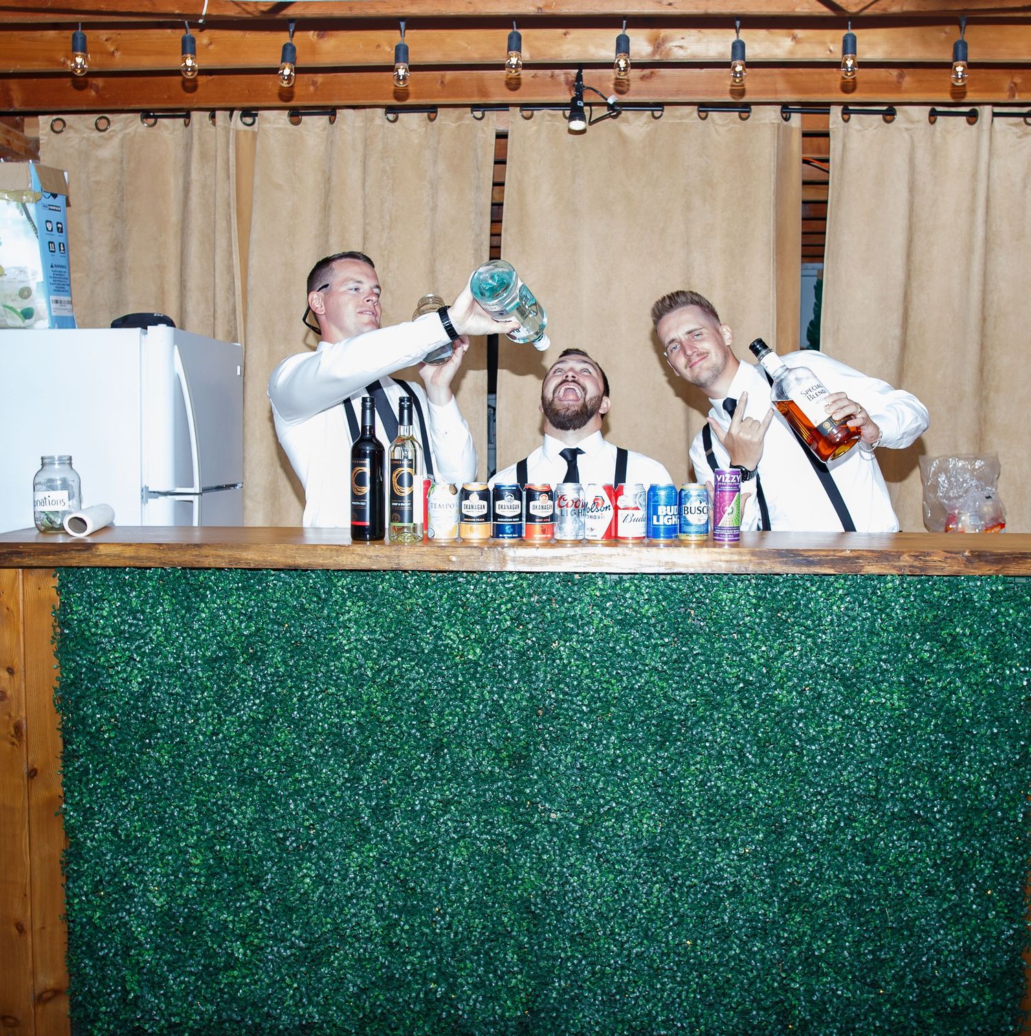 Wedding reception bar setup with wooden backdrop and artificial green hedge wall decoration.