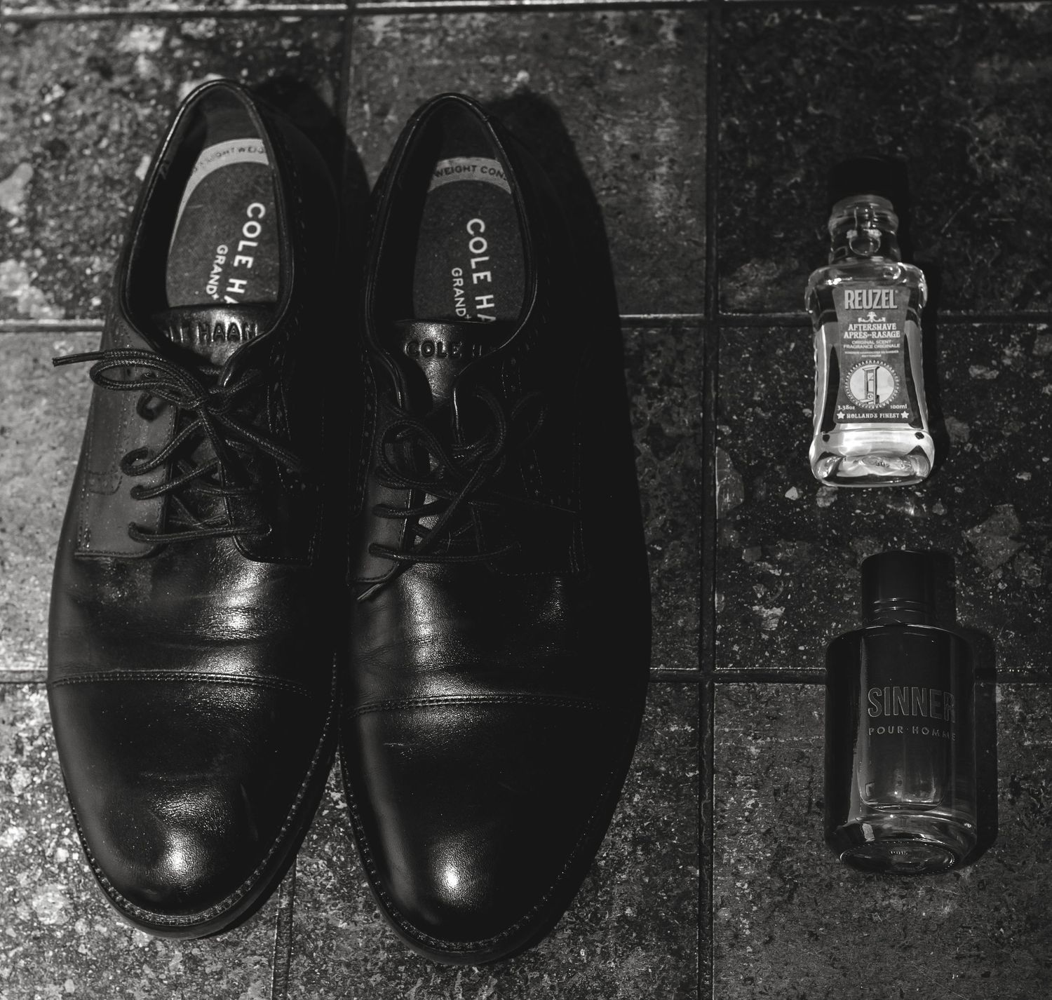 Black dress shoes and cologne bottle arranged on stone surface in monochrome.