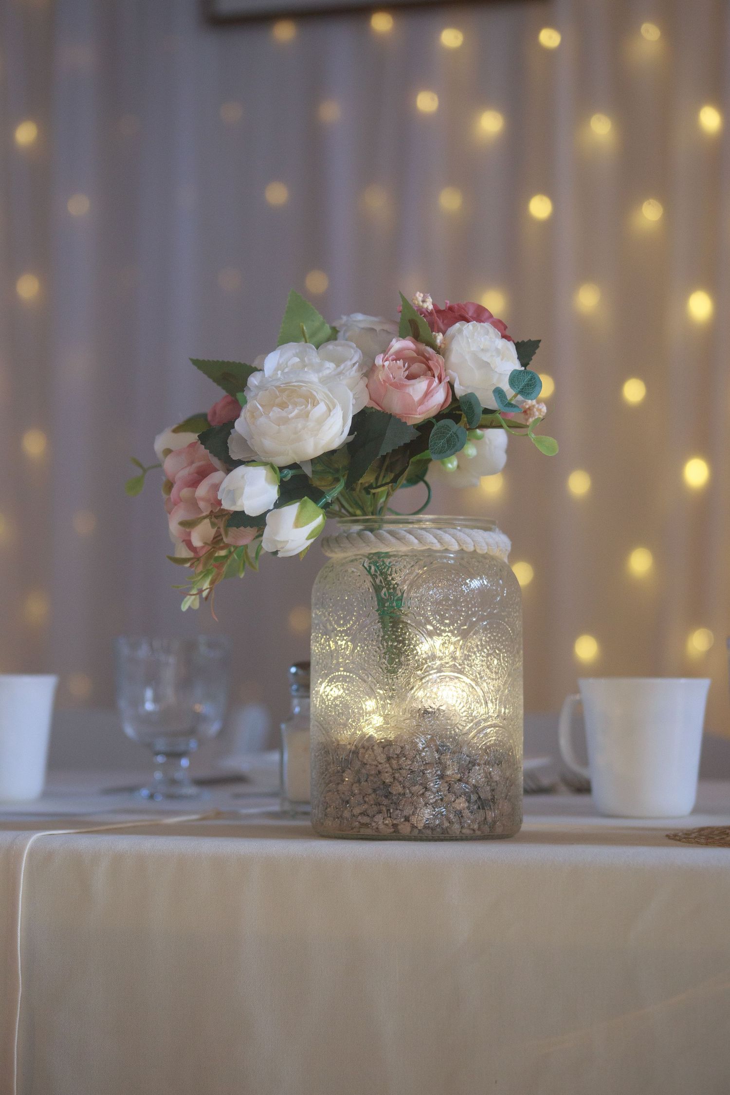 Mason jar centerpiece with pink roses and fairy lights against a bokeh background.