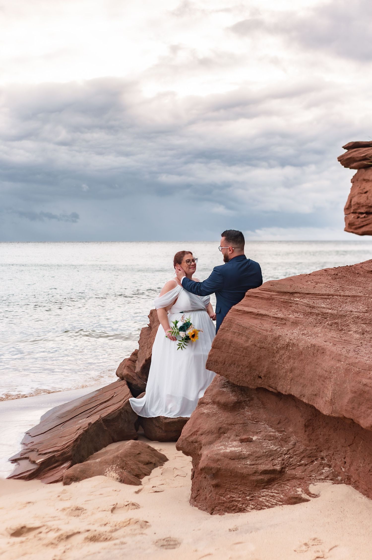 Wedding couple sitting on red rocky cliffs overlooking the ocean at sunset.