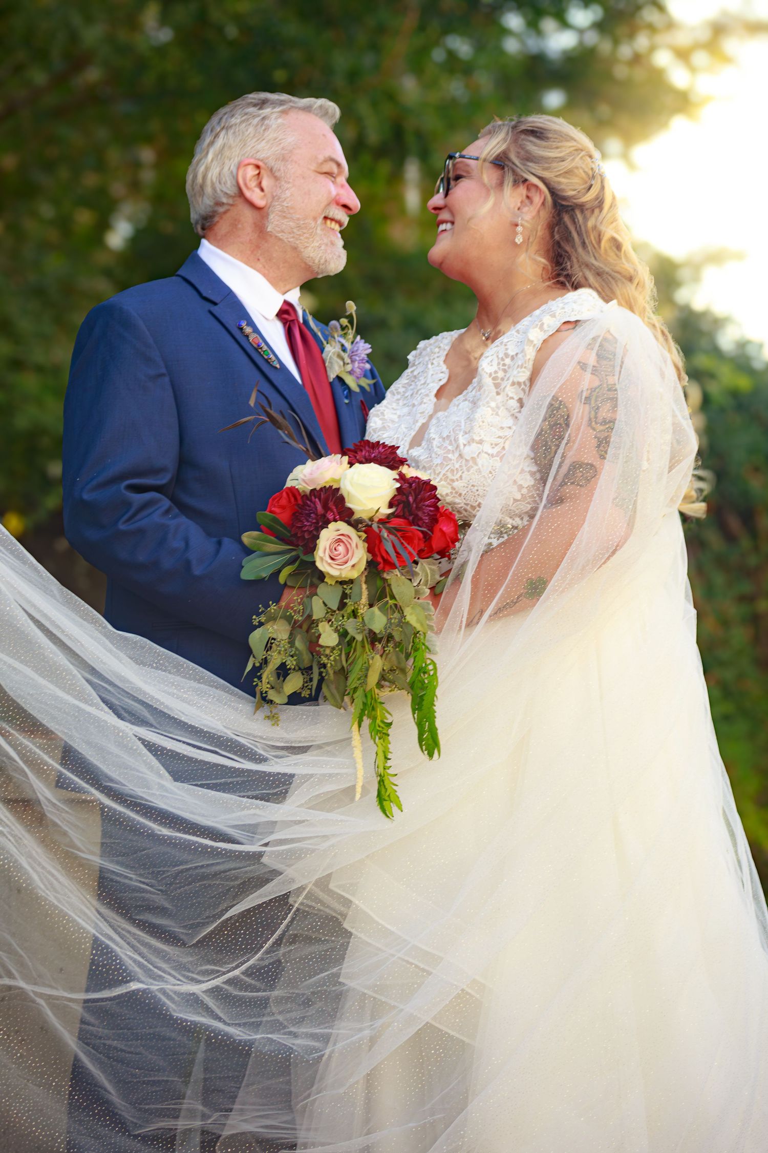 Wedding couple embraces outdoors with flowing veil and red bouquet in golden sunlight.