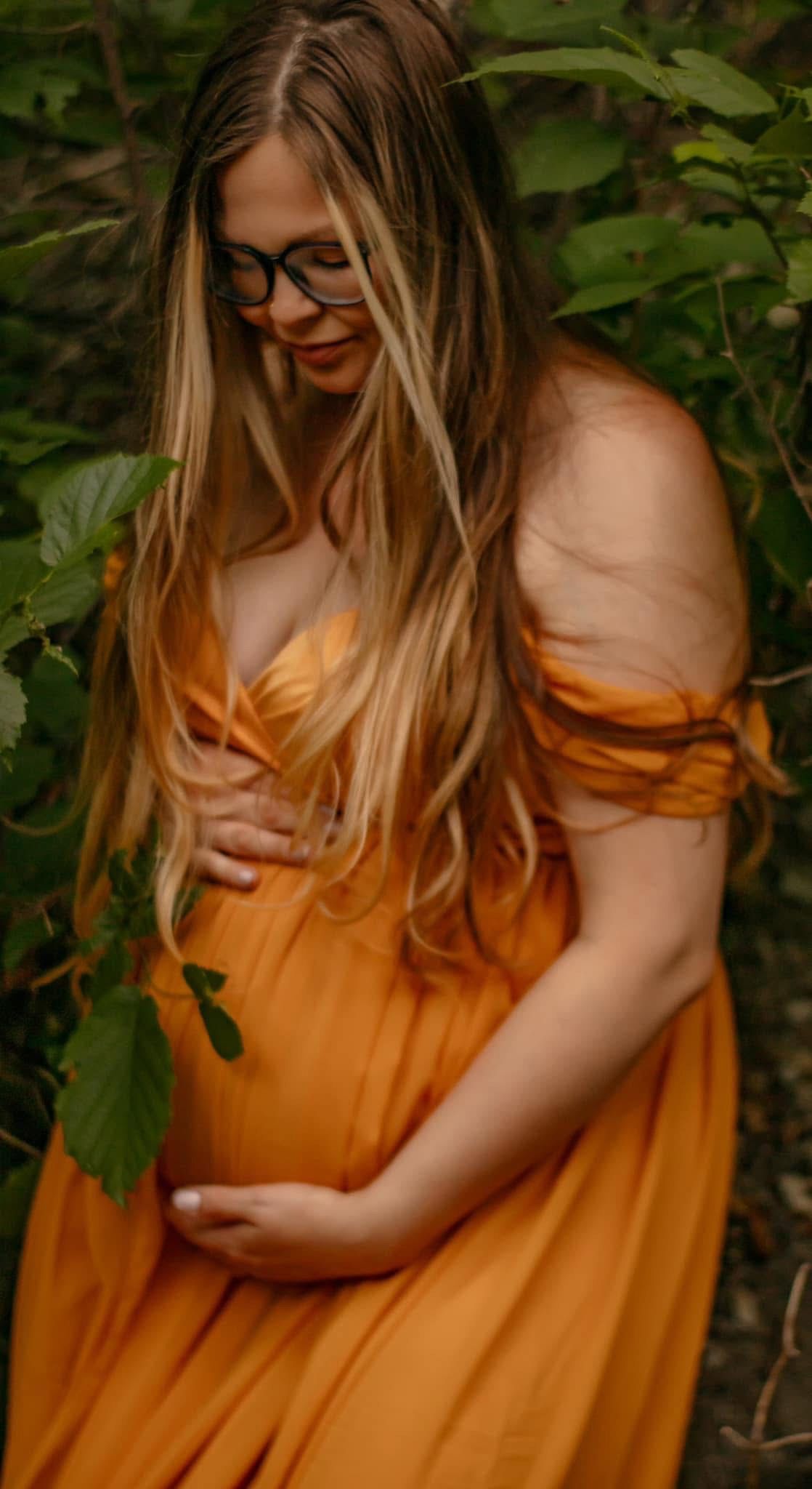 A dreamy autumn maternity portrait featuring a flowing orange dress among green foliage.