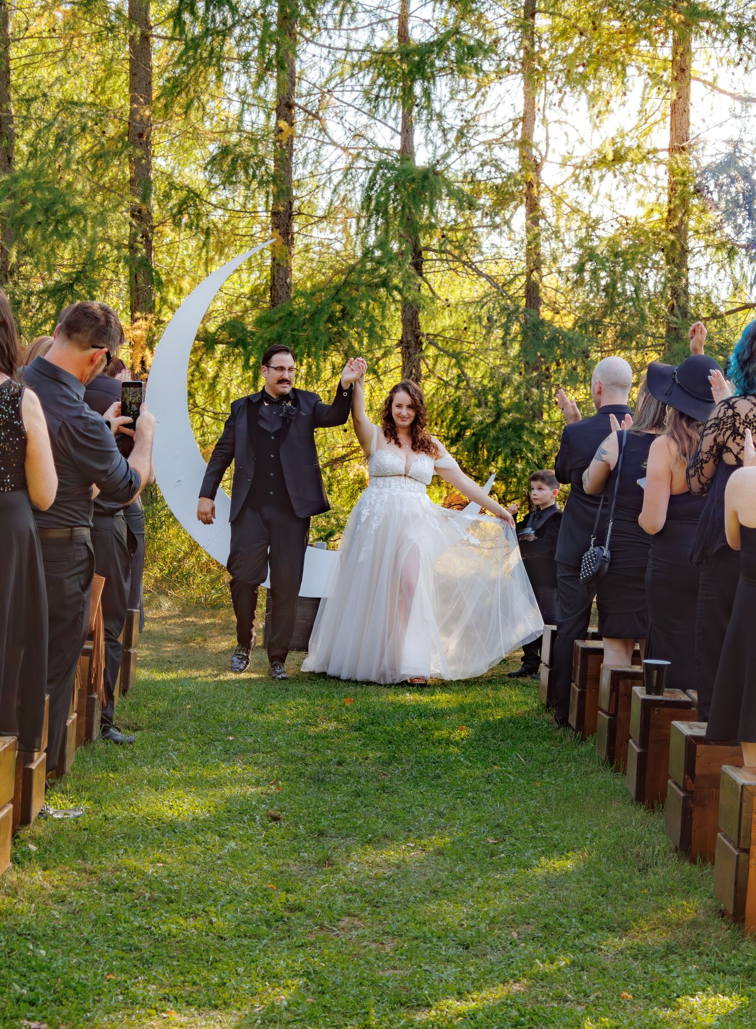 Wedding ceremony procession in a natural woodland setting with guests seated on rustic chairs.