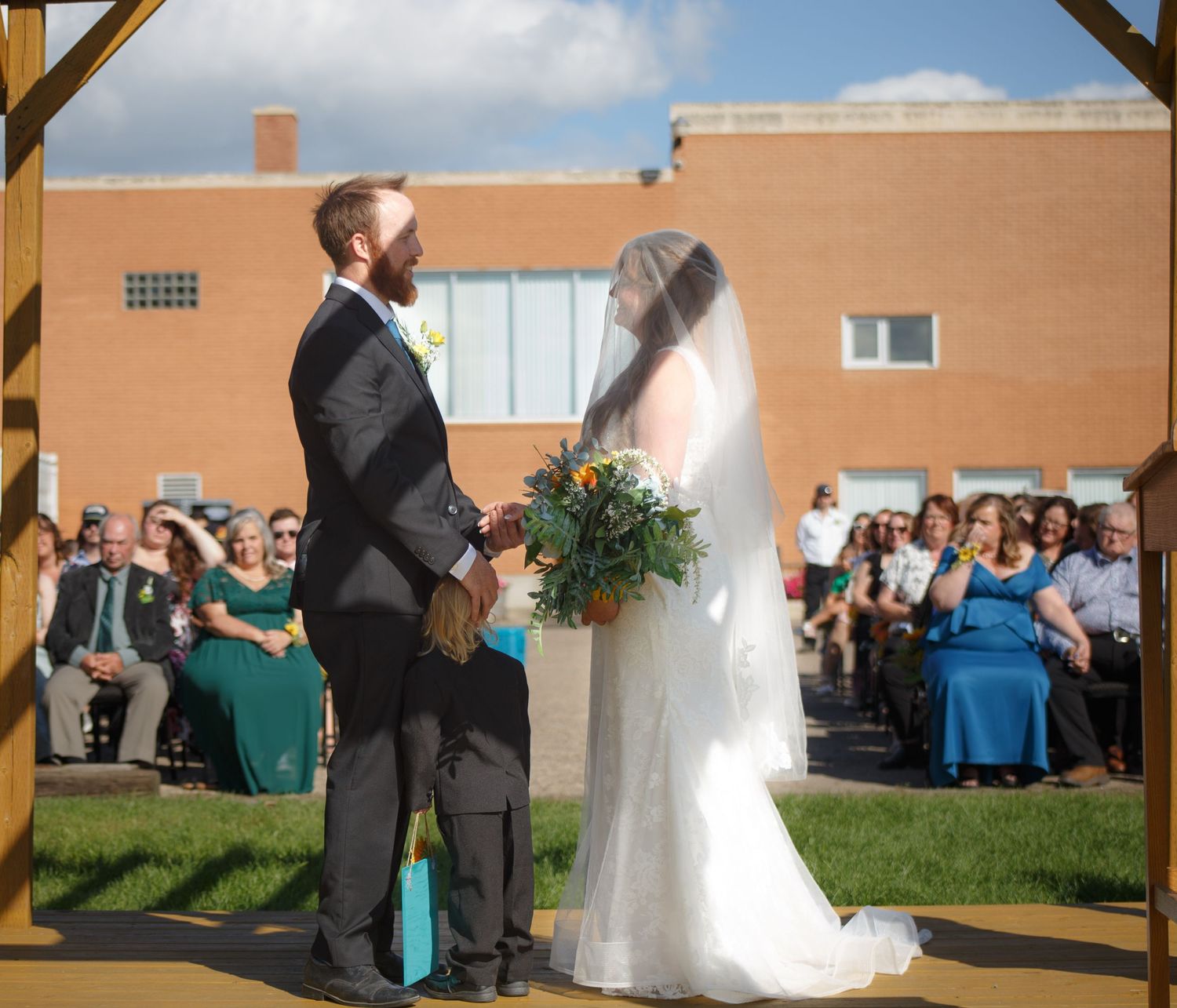 Outdoor wedding ceremony under wooden arch with guests seated on lawn.