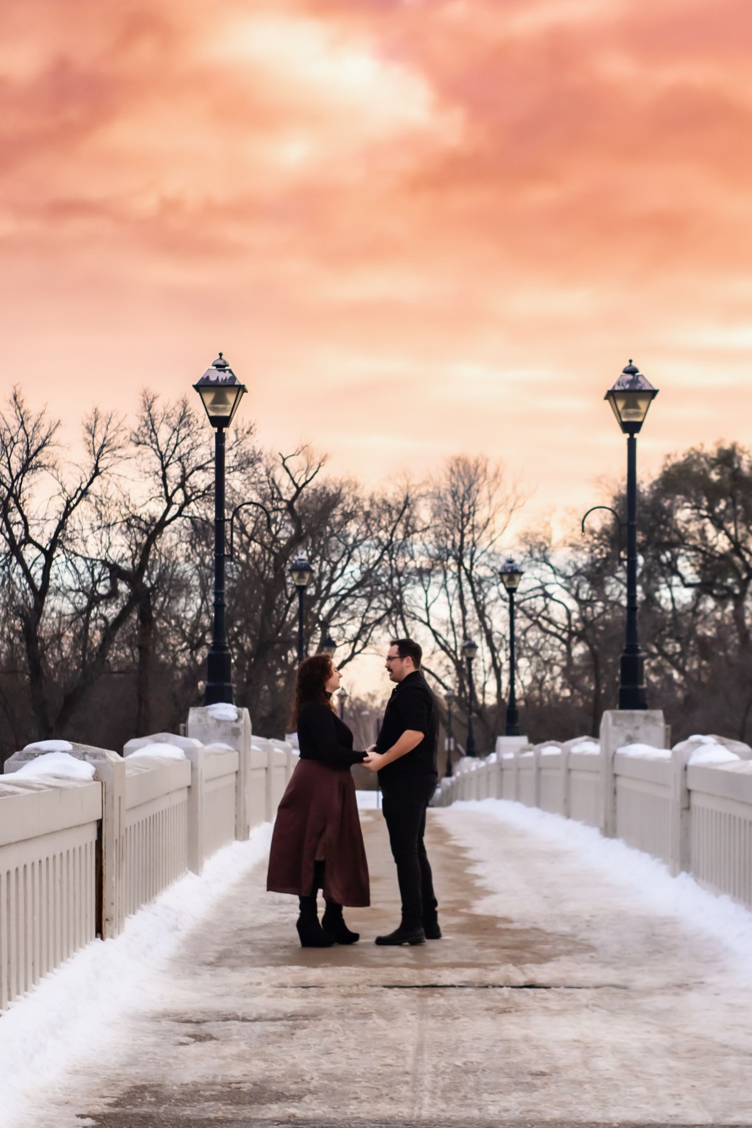 Couple standing on snowy path with white picket fence and vintage lamp posts at sunset.