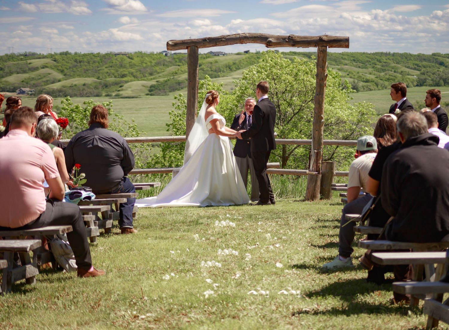 Outdoor wedding ceremony setup with wooden cross and guests seated on benches in a natural setting.
