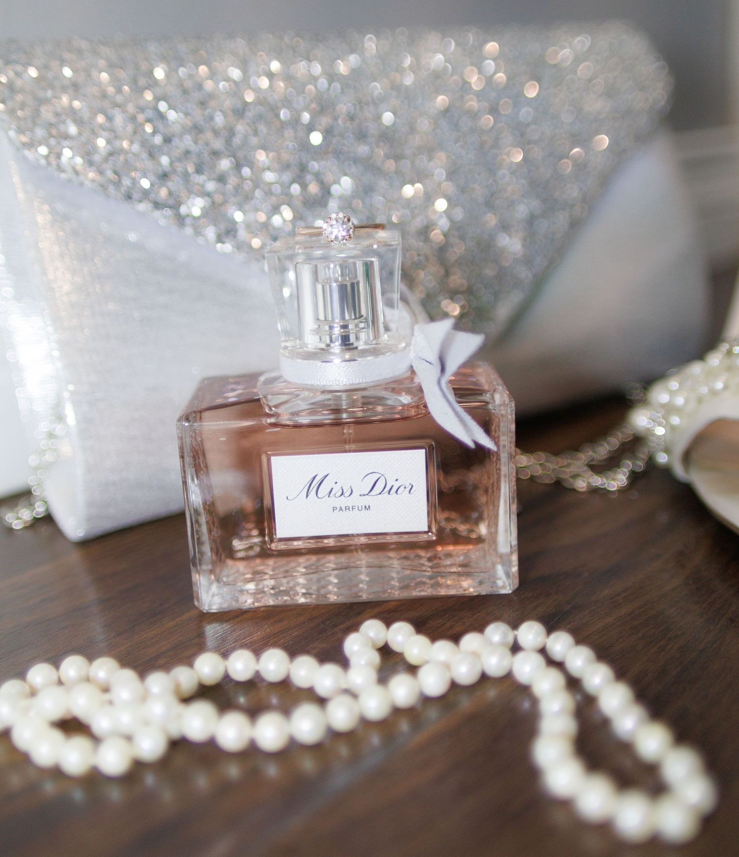Pearl necklace and perfume bottles arranged on wooden surface with sparkly backdrop.