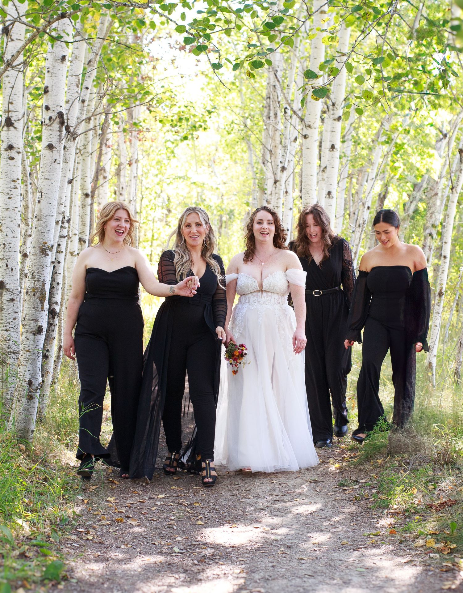 Wedding party poses in black attire among white birch trees on forest path.