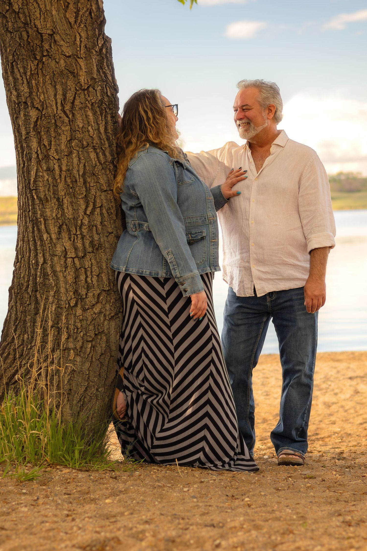 Couple in casual attire sharing tender moment against large tree trunk near waterfront.