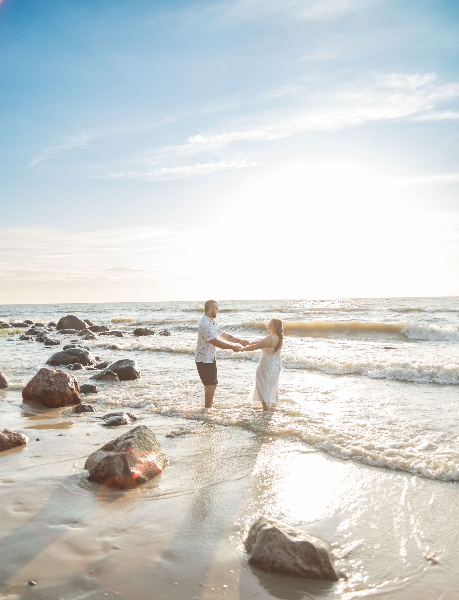 A couple holds hands while standing in ocean waves at sunset among coastal rocks.