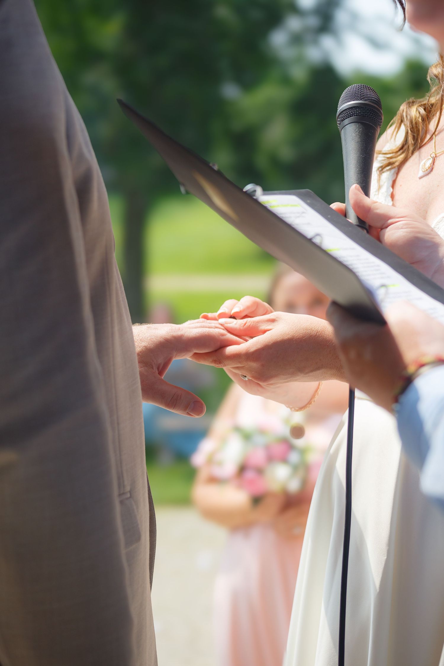 Wedding ceremony exchange of rings with officiant holding microphone outdoors.