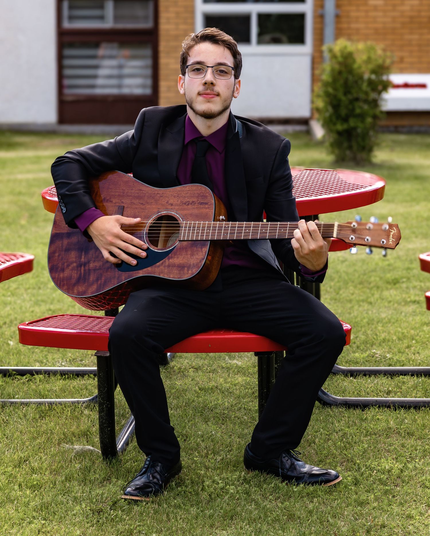Person in formal attire sitting on a red bench with an acoustic guitar outdoors.