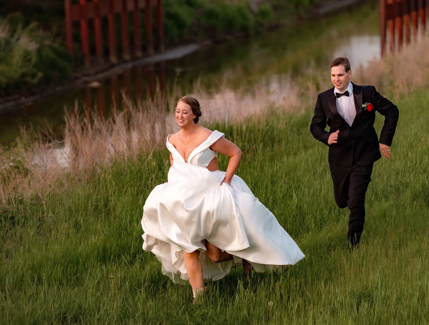 A couple runs playfully through a grassy field at sunset, with the bride's white dress flowing behind them.