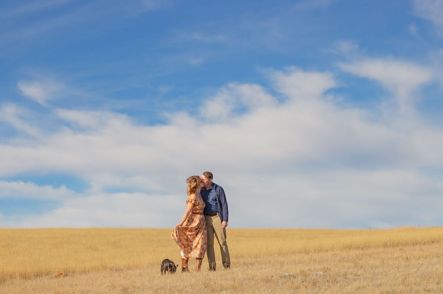 Couple walking together in golden wheat field under blue sky with white clouds.