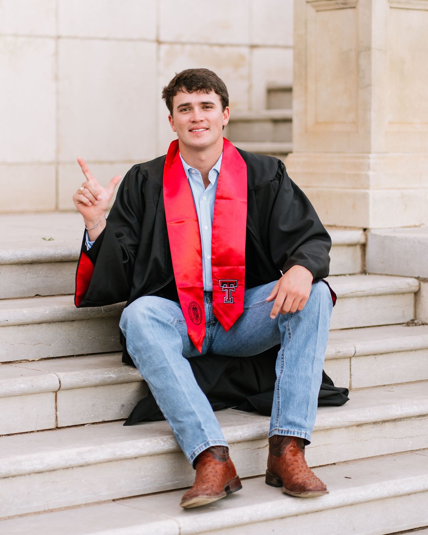 College graduate in black gown and red Texas Tech stole sits on stone steps, smiling and pointing upward.
