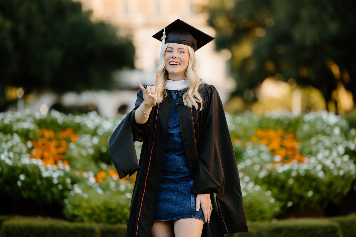 Graduate in black academic regalia poses in garden with flowers blooming.