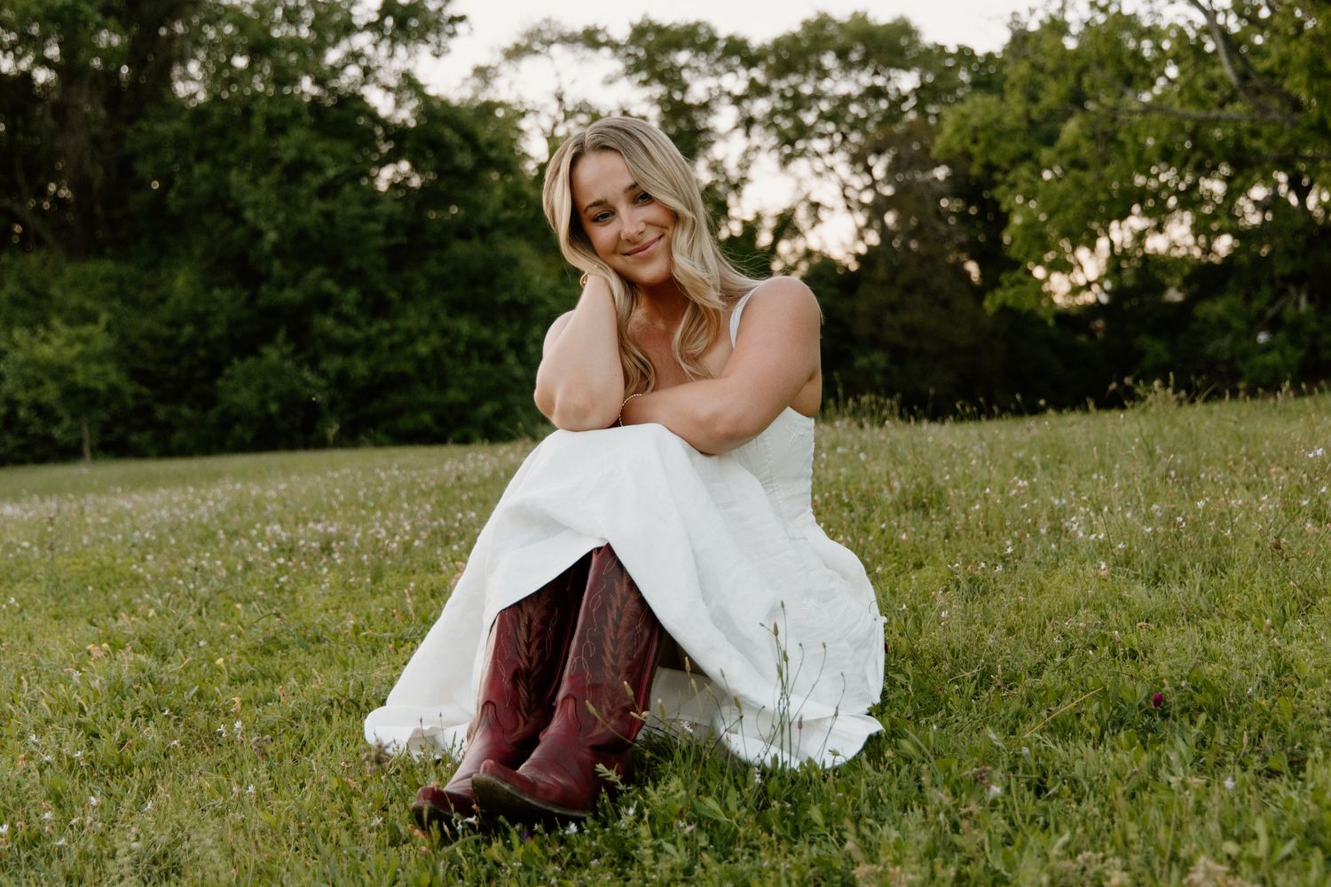 Woman in white dress and cowboy boots sitting in green field, smiling outdoors at sunset.