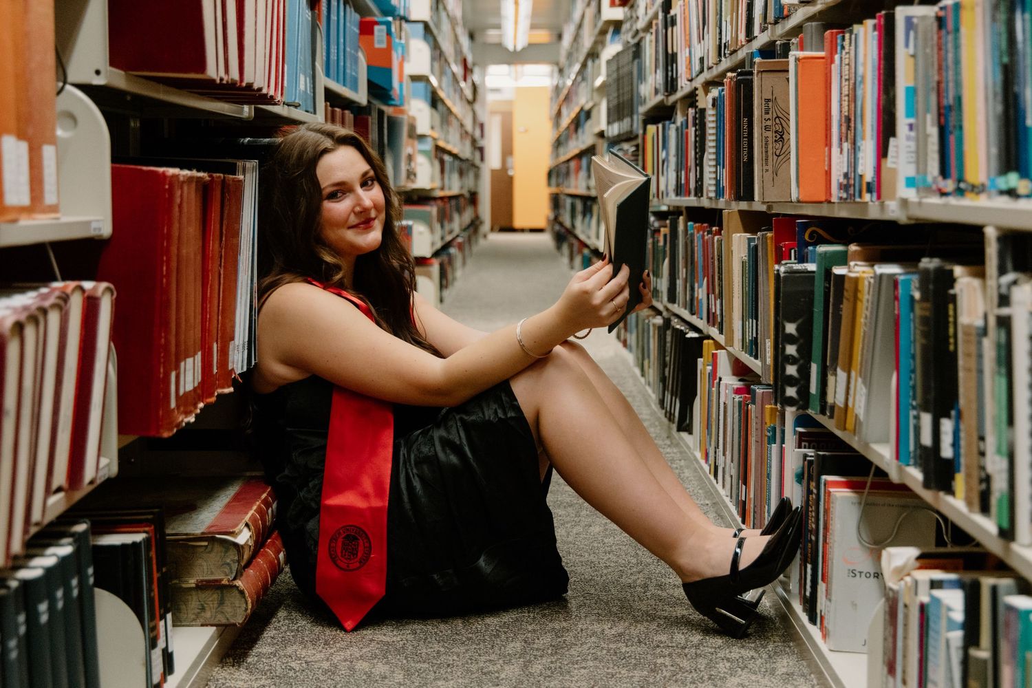 Graduate in cap and gown poses between library bookshelves for graduation photos.