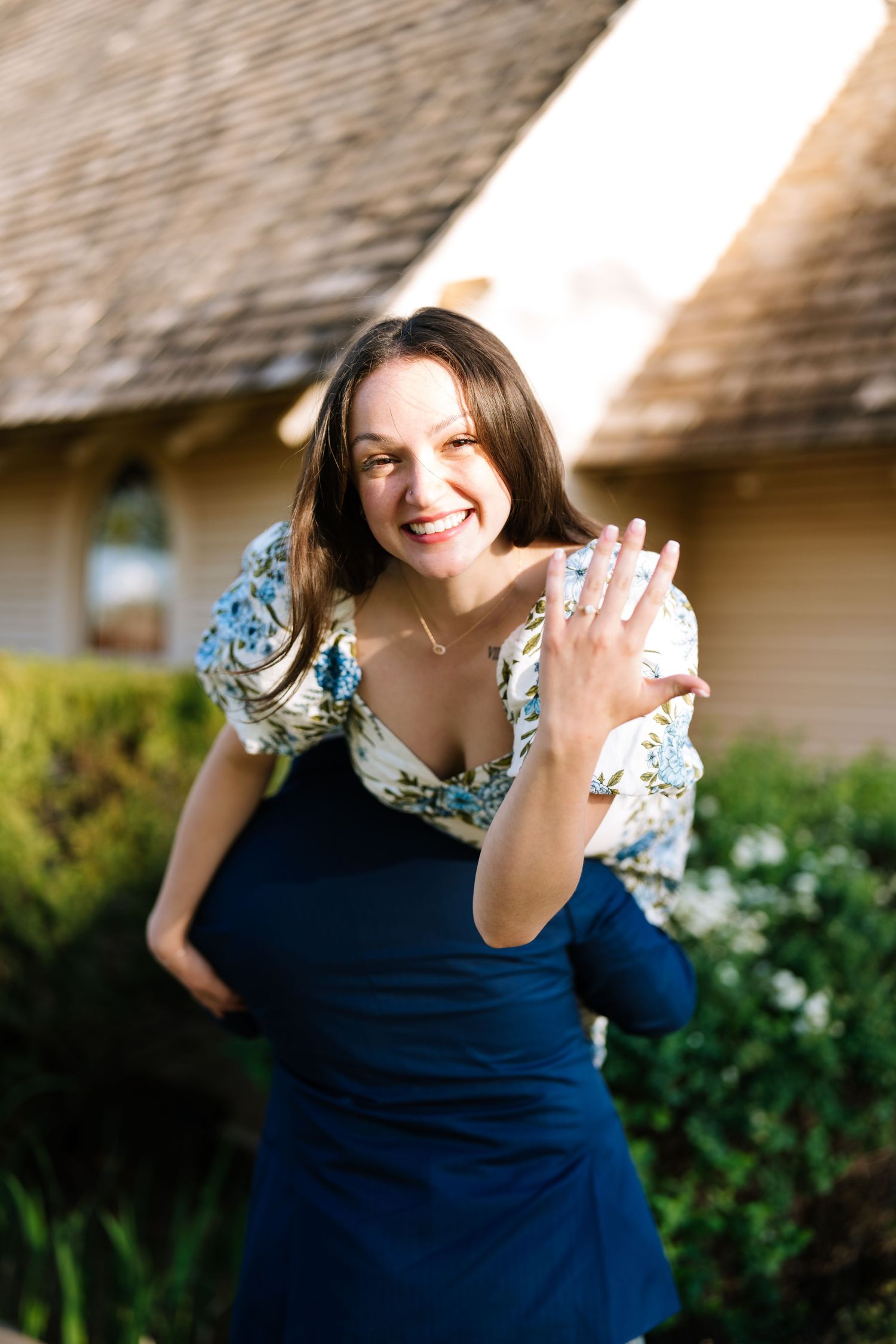 Smiling brunette woman in floral top and blue skirt showing off engagement ring outdoors near a house.