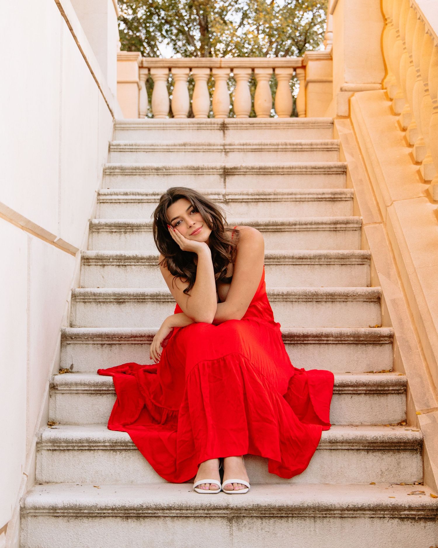 Woman in flowing red dress posing elegantly on white stone staircase.