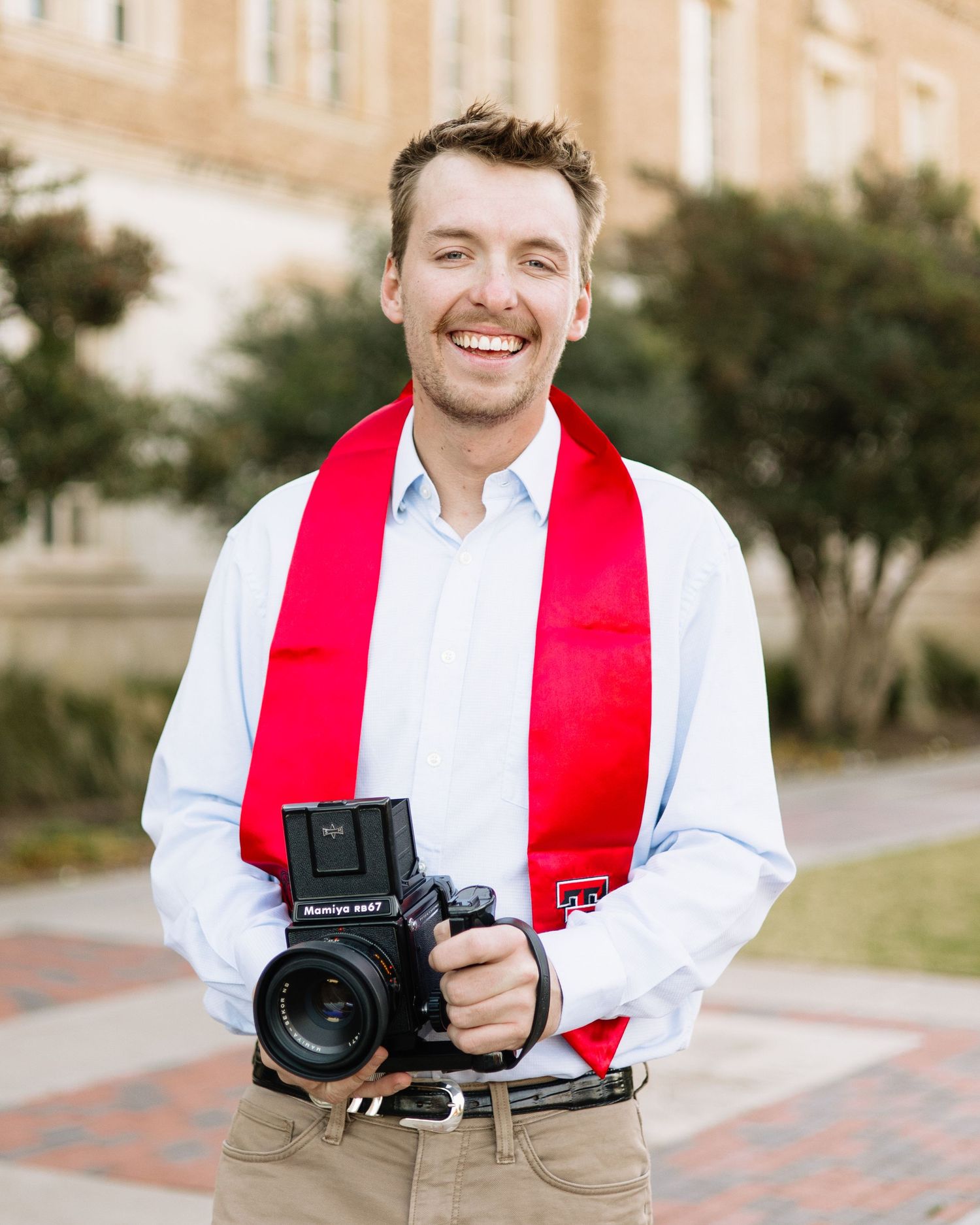 Graduate in red stole and white shirt holds professional camera during photo session.