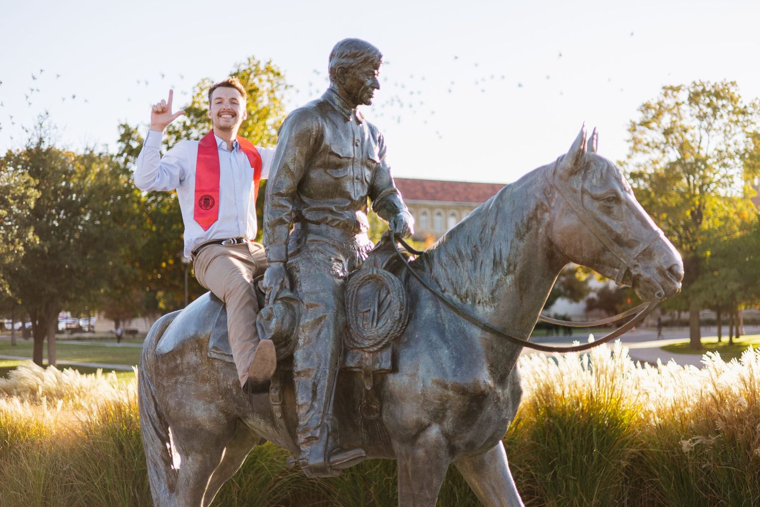 Student posing on a bronze equestrian statue on a sunny college campus, giving a thumbs up gesture.