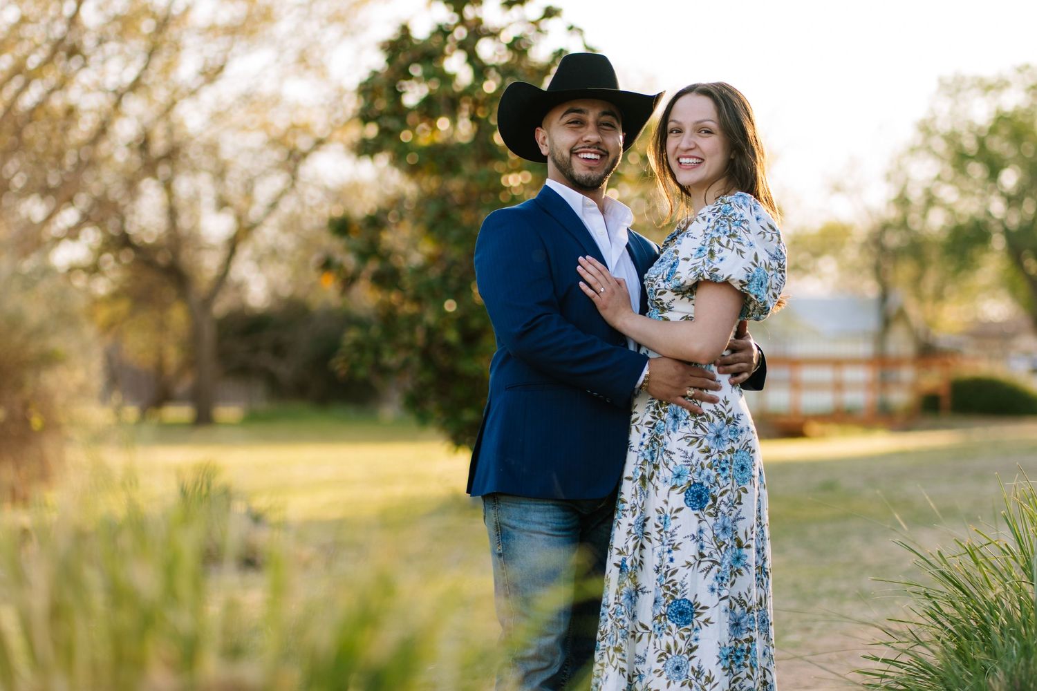 Couple embracing outdoors, man in blue blazer and cowboy hat, woman in floral dress, golden hour lighting.