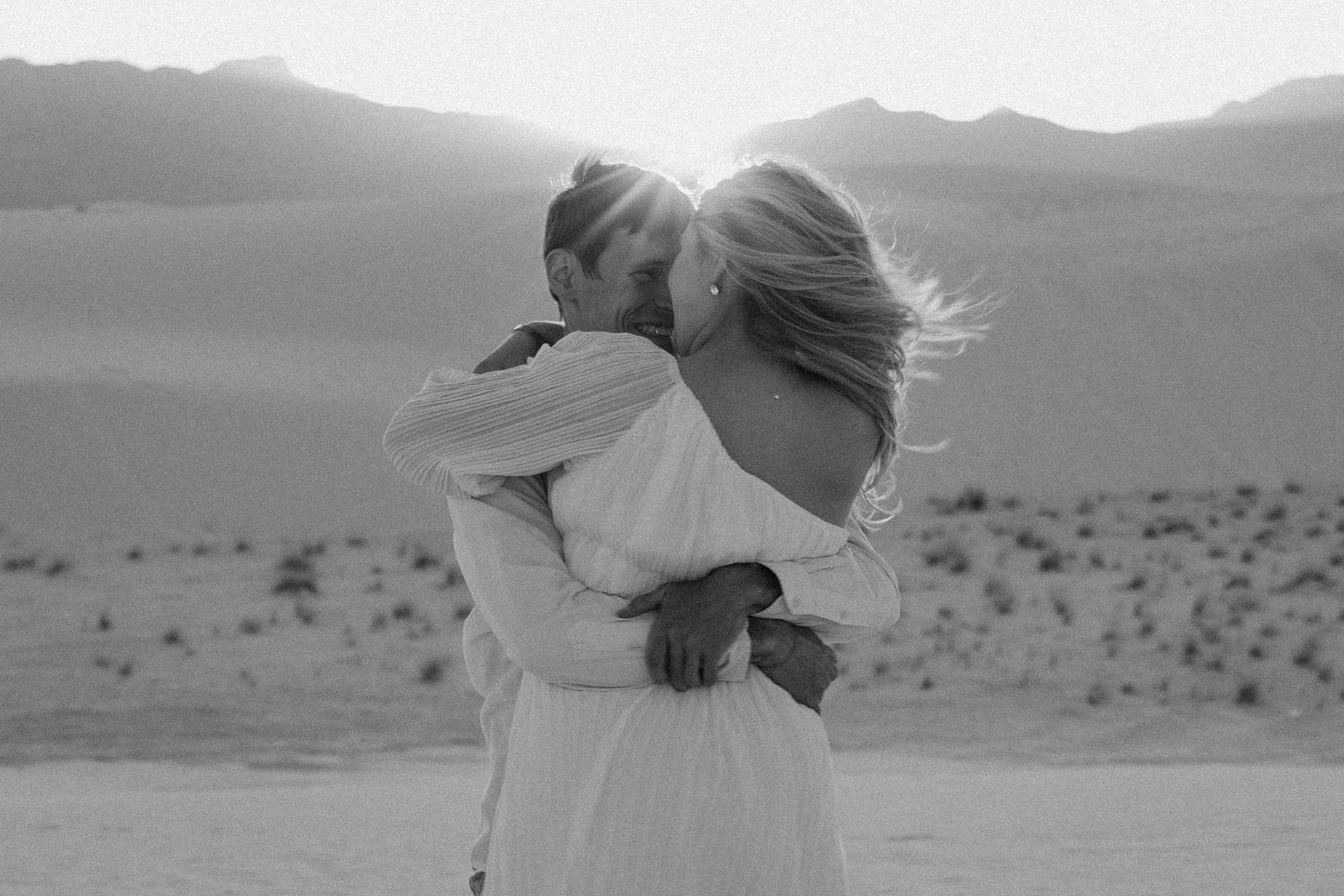 Couple embracing against desert mountain landscape at sunset in black and white.