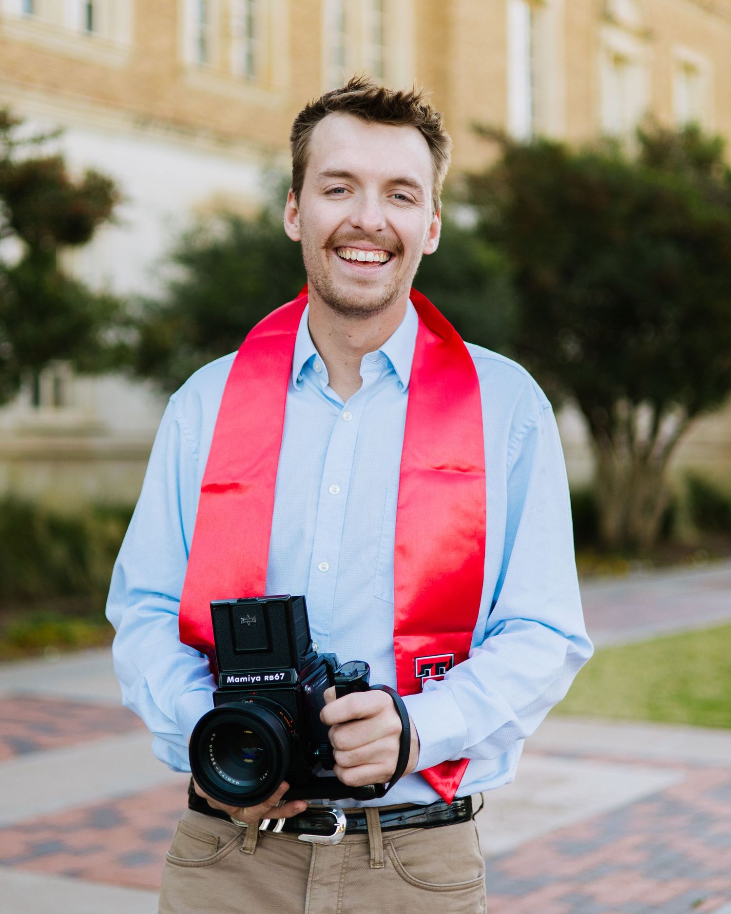 Professional photographer wearing a red stole and light blue shirt holds camera equipment outdoors.