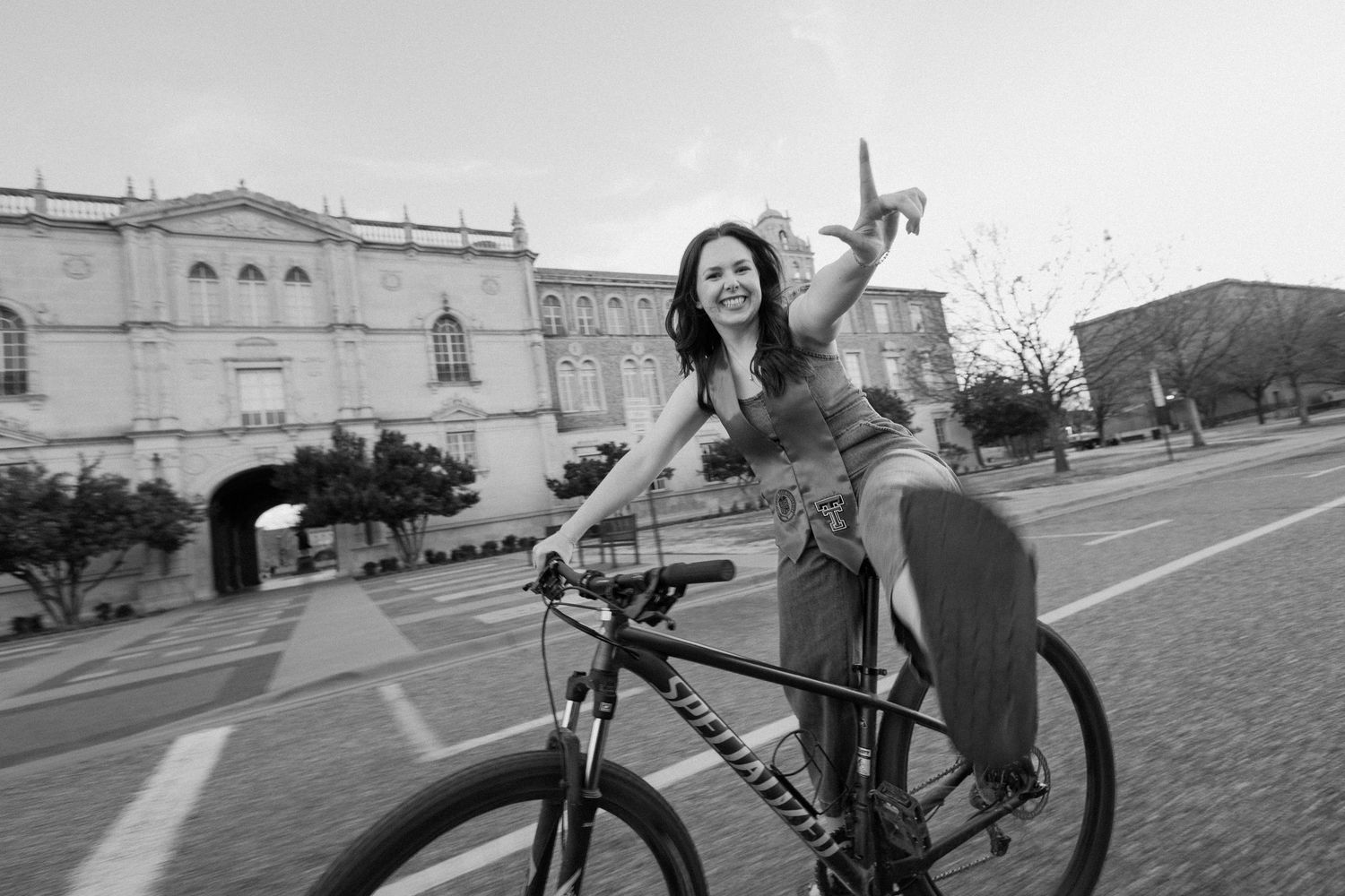 A smiling woman rides a mountain bike through a historic plaza, waving cheerfully in a black and white photo.