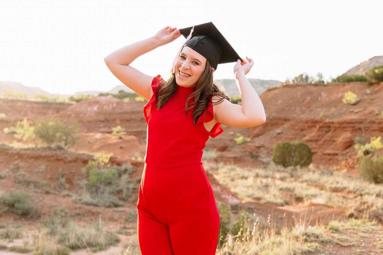 Graduate in red dress celebrating on desert landscape background.