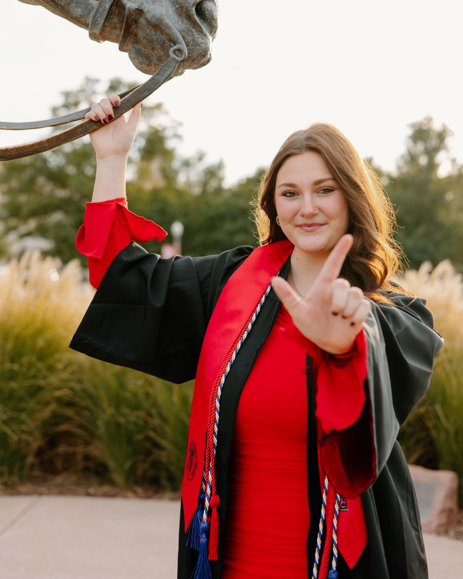 Graduate in black gown and red dress posing outdoors at sunset.