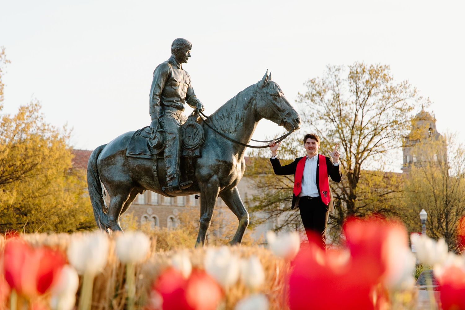 Person in red jacket poses beside bronze equestrian statue surrounded by red and white tulips in bloom.