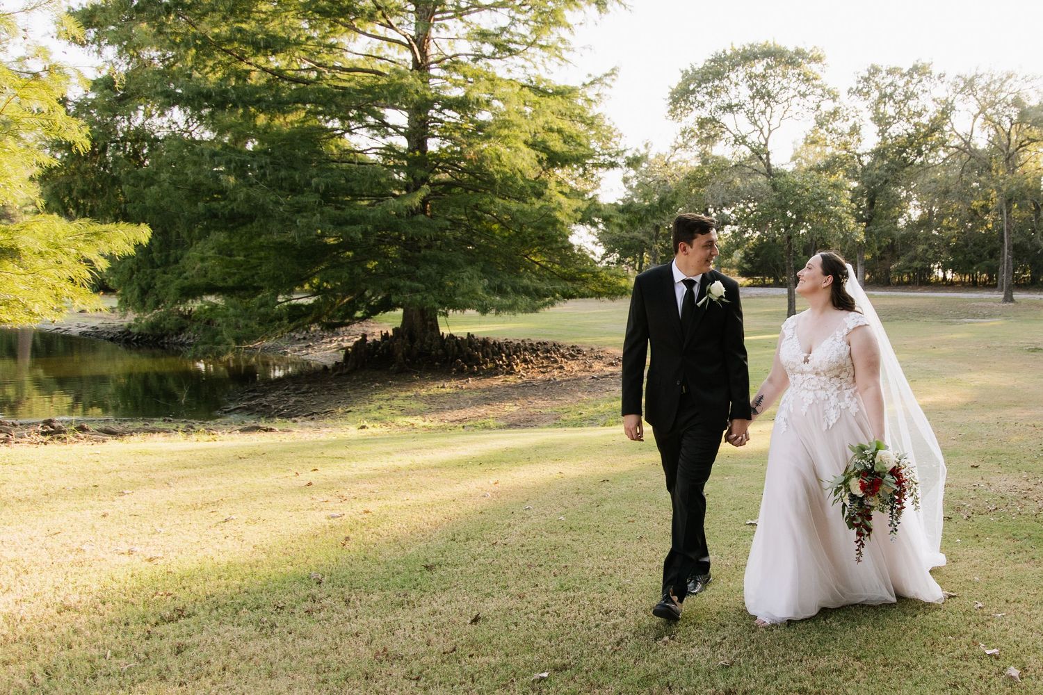 Bride and groom walk hand-in-hand across lush green lawn, surrounded by tall trees, sharing a romantic glance.