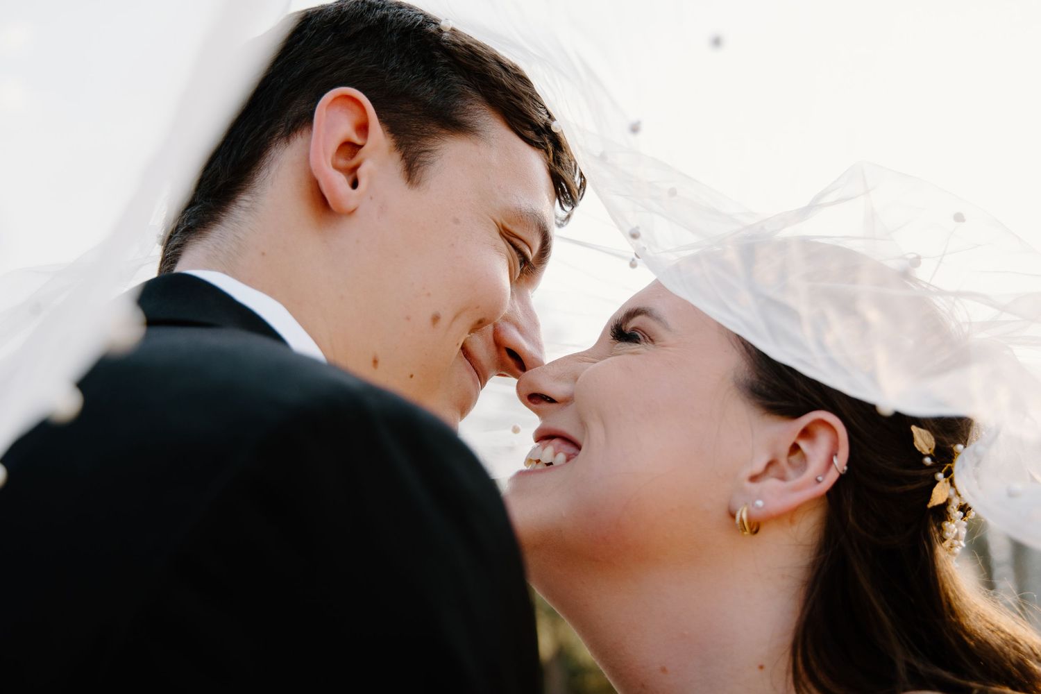A bride and groom share a romantic almost-kiss under a flowing white veil on their wedding day.