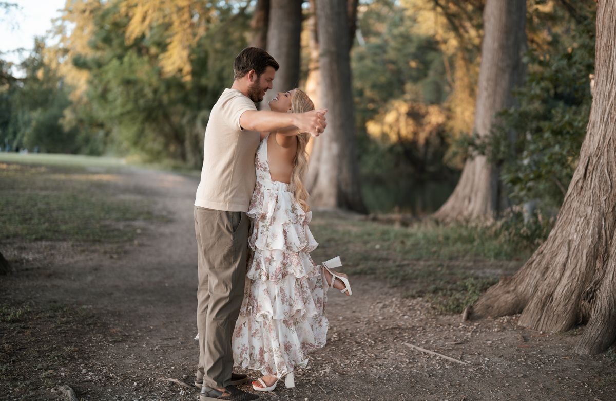 Romantic couple dancing outdoors among trees at sunset in casual attire.