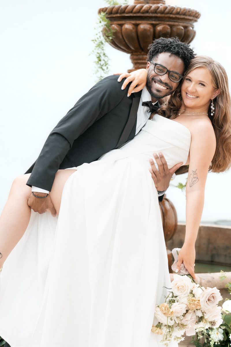 Happy couple in wedding attire share playful moment by decorative fountain.