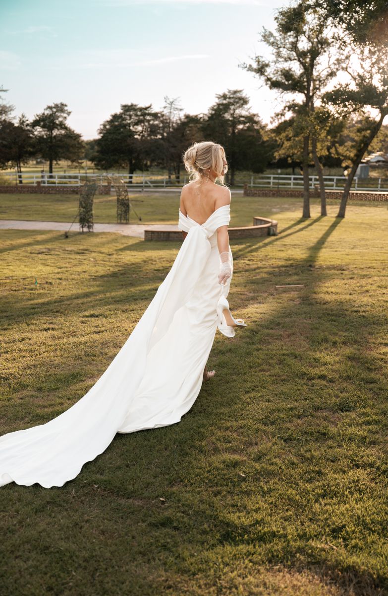 Bride in off-shoulder wedding gown walks across sunlit lawn with trailing dress.
