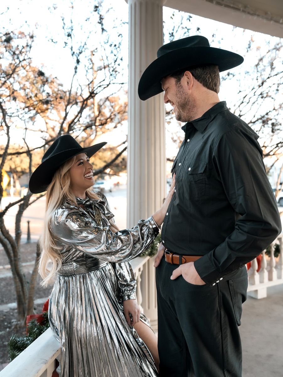 Couple in western attire shares intimate moment on porch with autumn trees in background.